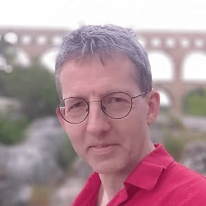 A person with short gray hair and glasses, wearing a red shirt, standing in front of a stone structure with arches in the background.