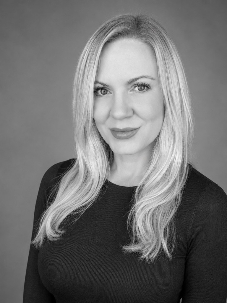 Black and white portrait of a woman with long blond hair, wearing a fitted long-sleeve top, smiling at the camera.