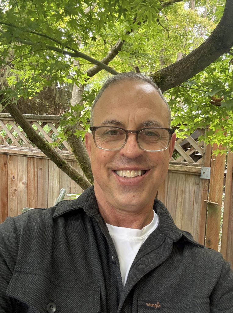 A smiling man with short hair and glasses, wearing a dark jacket, poses for a selfie in a garden with green leaves and wooden fencing in the background.