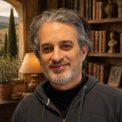 A middle-aged man with gray hair and a beard, wearing a dark hoodie, standing in a cozy room with bookshelves and warm lighting.